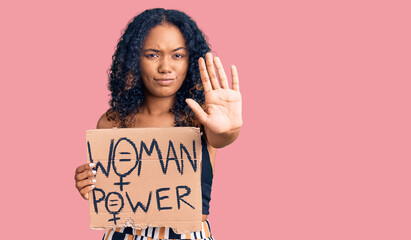 Young african american girl holding woman power banner with open hand doing stop sign with serious...