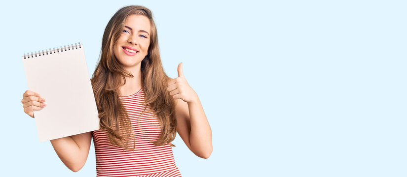 Beautiful caucasian young woman holding notebook smiling happy and positive, thumb up doing excellent and approval sign