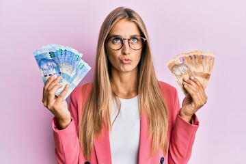 Young blonde woman wearing business style holding south african rands banknotes making fish face with mouth and squinting eyes, crazy and comical.