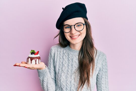 Young beautiful caucasian girl wearing french beret holding cake looking positive and happy standing and smiling with a confident smile showing teeth
