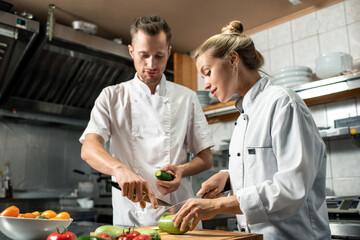 Young confident professional chef with knife and cucumber consulting his trainee