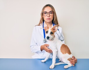 Young beautiful blonde veterinarian woman checking dog health relaxed with serious expression on...