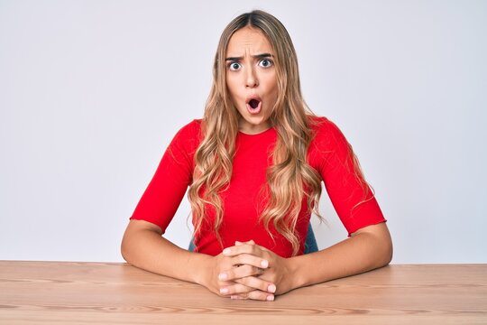 Young beautiful blonde woman wearing casual clothes sitting on the table scared and amazed with open mouth for surprise, disbelief face