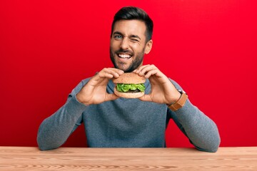 Handsome hispanic man eating a tasty classic burger winking looking at the camera with sexy...