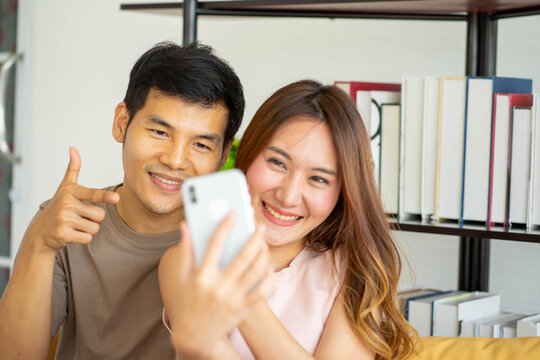 portrait of Asian man giving ring engagement wedding to happy exciting woman sitting in couch with cellular mobile phone camera selfie recorind picture  ar home.