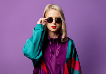 Style blonde in 80s windbreaker and roud sunglasses on purple background