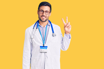 Young hispanic man wearing doctor uniform and stethoscope smiling looking to the camera showing fingers doing victory sign. number two.