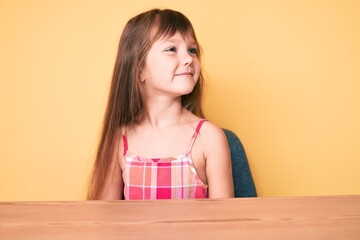 Little caucasian kid girl with long hair wearing casual clothes sitting on the table looking to side, relax profile pose with natural face and confident smile.