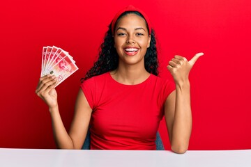 Young african american girl holding israel shekels sitting on the table pointing thumb up to the...