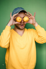Young man with a beard in a cap on his head with lemons on his eyes on a green background. Portrait of a guy in a yellow sweater