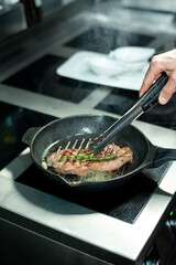 Hand of chef frying piece of beef and adding fresh aromatic herbs to the meat