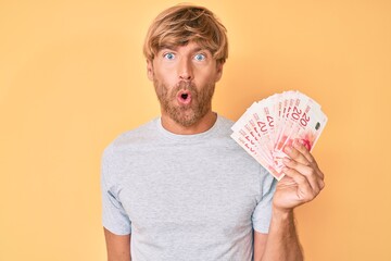 Young blond man holding israeli shekels banknotes scared and amazed with open mouth for surprise, disbelief face