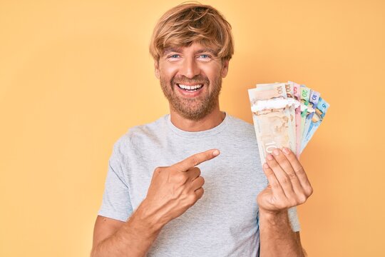 Young blond man holding canadian dollars smiling happy pointing with hand and finger