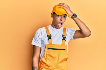 Young handsome african american man wearing handyman uniform over yellow background surprised with hand on head for mistake, remember error. forgot, bad memory concept.