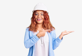 Young latin woman wearing architect hardhat amazed and smiling to the camera while presenting with hand and pointing with finger.