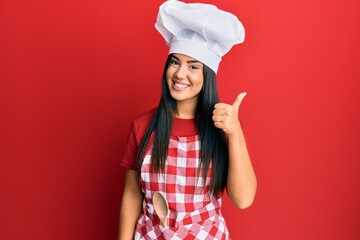 Young beautiful hispanic girl wearing baker uniform and cook hat smiling happy and positive, thumb up doing excellent and approval sign