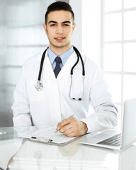 Arab doctor man is using laptop computer while filling up medication history records form at the glass desk in r clinic. Medicine concept