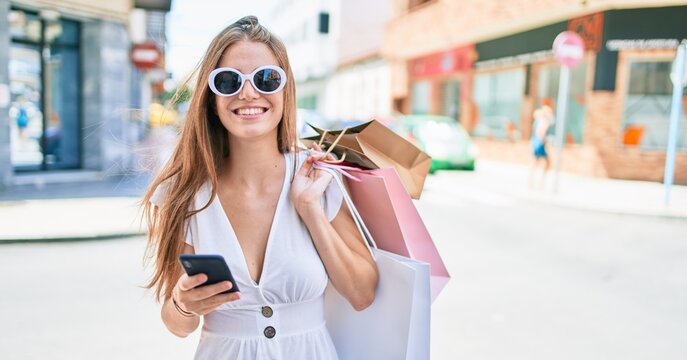 Young blonde woman smiling happy holding shopping bags and using smartphone at street of city