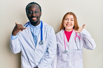 Young interracial couple wearing doctor uniform and stethoscope smiling doing phone gesture with...
