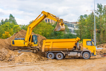 Yellow truck and excavator load soil with sand during road construction in urban area.