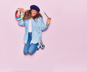 Young beautiful curly artist woman with tattoo wearing french beret smiling happy. Jumping with smile on face painting using palette and paintbrush over isolated pink background