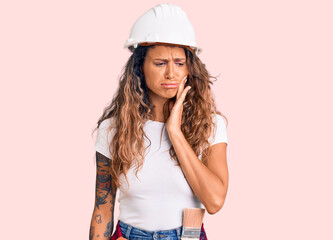 Young hispanic woman with tattoo wearing hardhat and builder clothes touching mouth with hand with painful expression because of toothache or dental illness on teeth. dentist