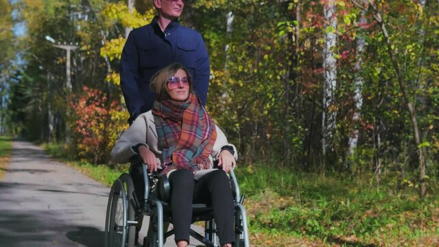 Young caucasian woman sitting on a wheelchair and caucasian young man walks in beautiful autumn park . Man takes care of handicapped woman outdoors