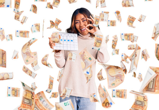 Young beautiful mixed race woman holding travel calendar doing ok sign with fingers, smiling friendly gesturing excellent symbol