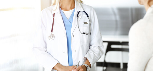 Unknown woman-doctor communicating with female patient in sunny clinic. Physician checks medical history record and exam results, close-up