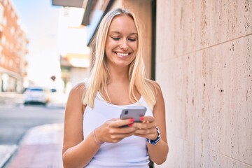 Young blonde girl smiling happy using smartphone at the city.