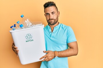 Handsome man with beard holding recycling wastebasket with plastic bottles thinking attitude and sober expression looking self confident