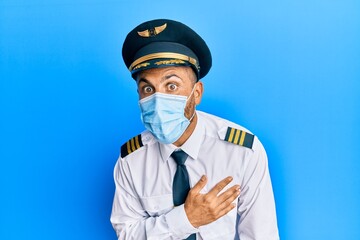 Handsome man with beard wearing airplane pilot uniform wearing safety mask scared and amazed with open mouth for surprise, disbelief face