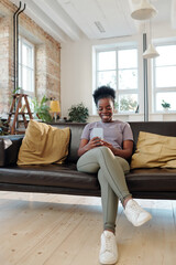 Obraz premium Restful young African woman with smartphone sitting on black leather couch