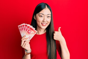 Young chinese woman holding 20 israel shekels banknotes smiling happy and positive, thumb up doing...