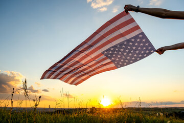 Human hands holding waving american national flag in field at sunset.