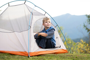 Happy boy resting alone in a tourist tent at mountain campsite enjoying view of beautiful summer nature. Hiking and active way of life concept.