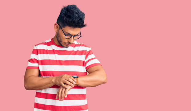 Handsome latin american young man wearing casual clothes and glasses checking the time on wrist watch, relaxed and confident