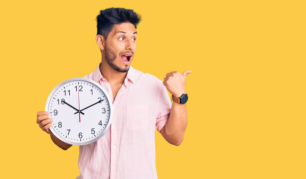 Handsome latin american young man holding big clock pointing thumb up to the side smiling happy with open mouth