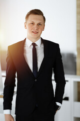 Cheerful businessman standing and looking at camera in sunny office. Headshot of young entrepreneur