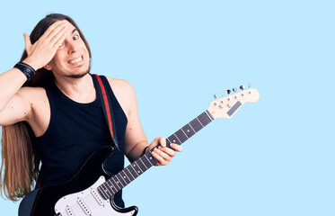 Obraz premium Young adult man with long hair playing electric guitar stressed and frustrated with hand on head, surprised and angry face