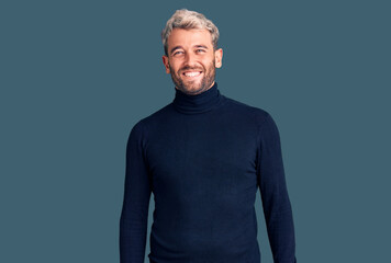 Obraz premium Young handsome blond man wearing casual turtleneck sweater looking positive and happy standing and smiling with a confident smile showing teeth