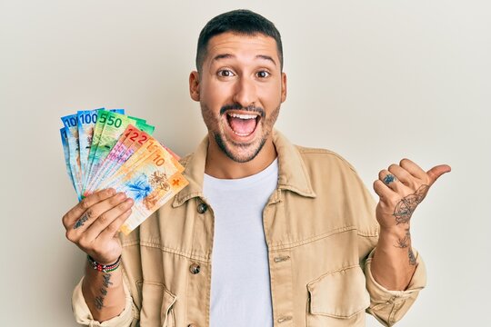 Handsome man with tattoos holding swiss franc banknotes pointing thumb up to the side smiling happy with open mouth
