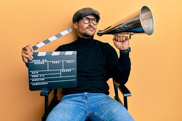 Handsome man with tattoos holding video film clapboard and louder smiling looking to the side and...