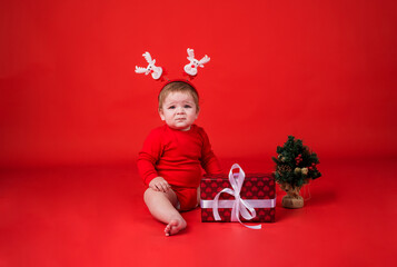 a beautiful baby girl in a Christmas headband with reindeer sits with a small Christmas tree and a gift box on a red background with space for text