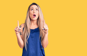 Young beautiful blonde woman wearing casual sleeveless t-shirt amazed and surprised looking up and...