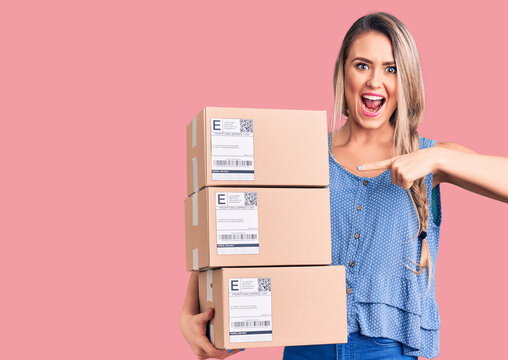 Young beautiful blonde woman holding delivery boxes smiling happy pointing with hand and finger