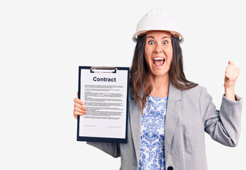 Young beautiful brunette woman wearing architect hardhat holding contract screaming proud, celebrating victory and success very excited with raised arms