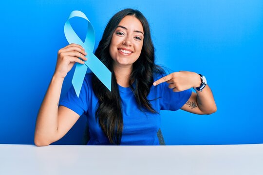 Beautiful brunette young woman holding blue ribbon pointing finger to one self smiling happy and proud