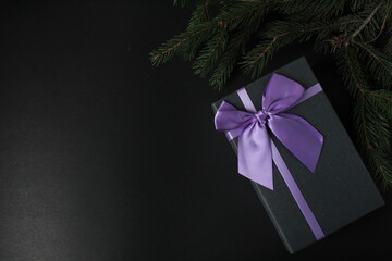 new year's Christmas background a branch of spruce pine trees on a black background and a gift box black gift box with lilac purple fuchsia ribbon and bow minimalism top view from copy space