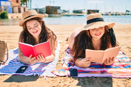 Two plus size overweight sisters twins women happy relaxing reading a book at the beach on summer holidays
